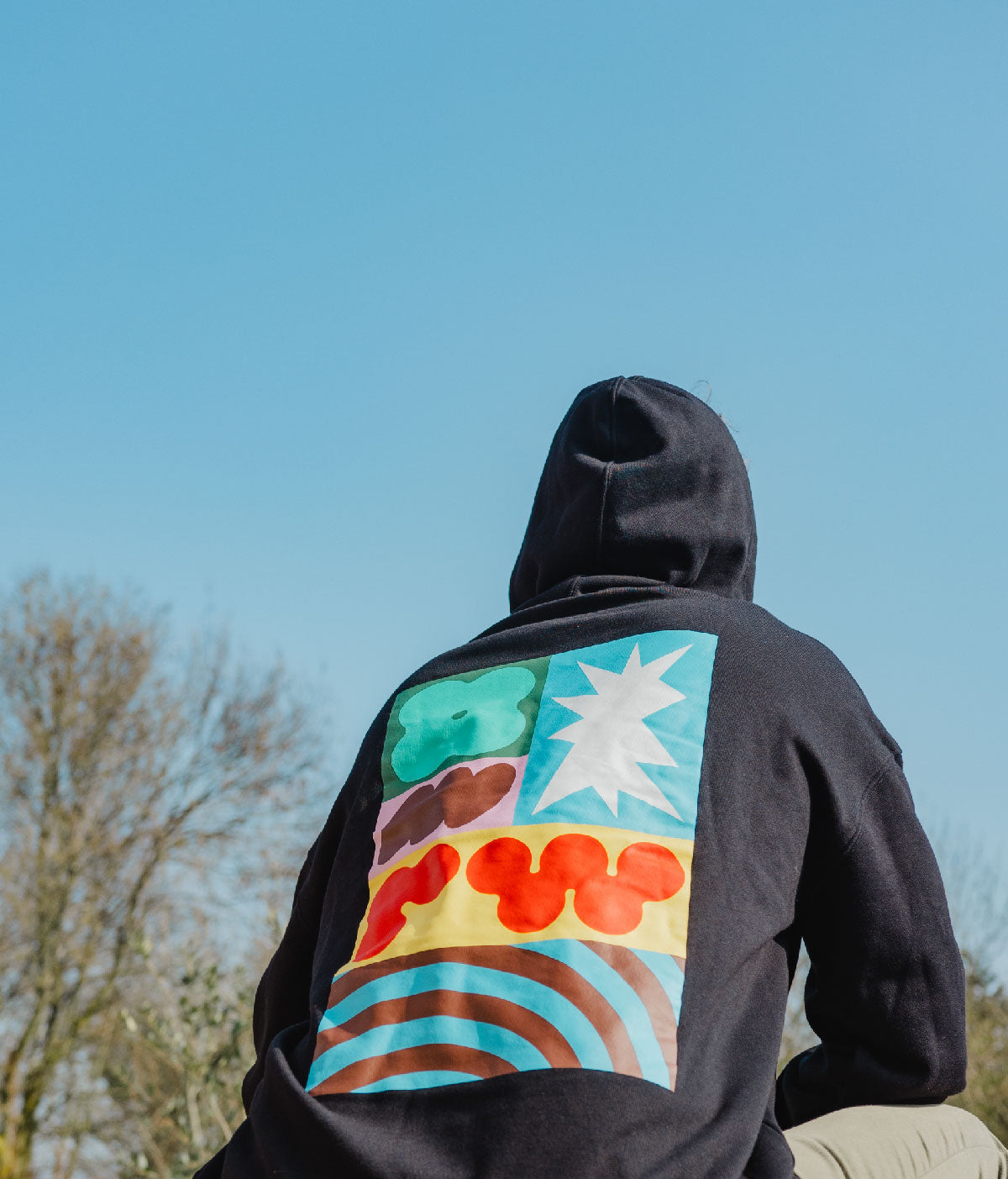 Person wearing a black hoodie with a colorful back design against a blue sky.
