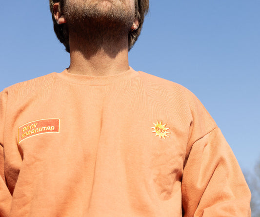 Person wearing an orange sweatshirt with a logo and sun design against a blue sky.