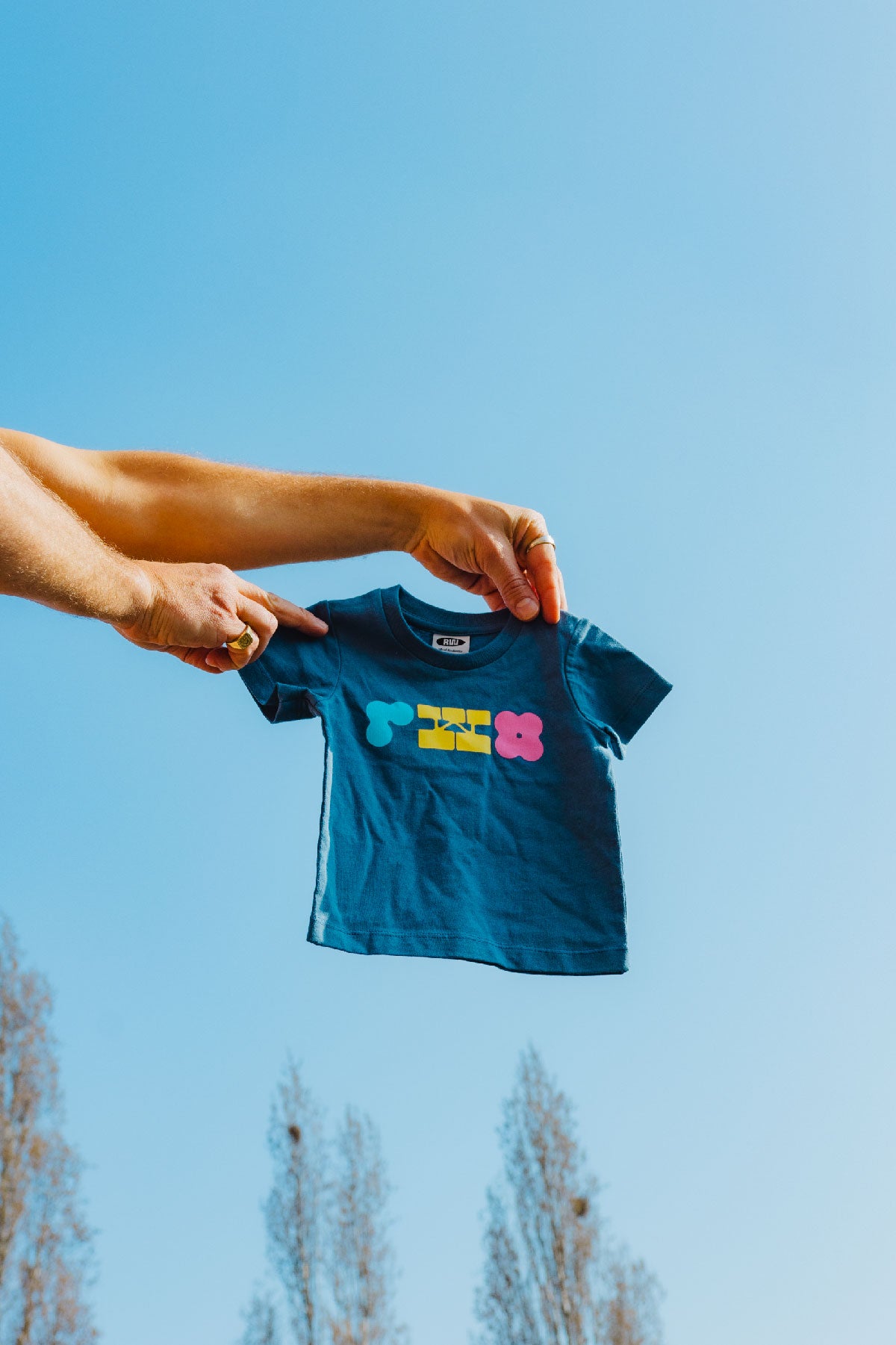 Person holding a blue t-shirt with colorful text against a clear blue sky.